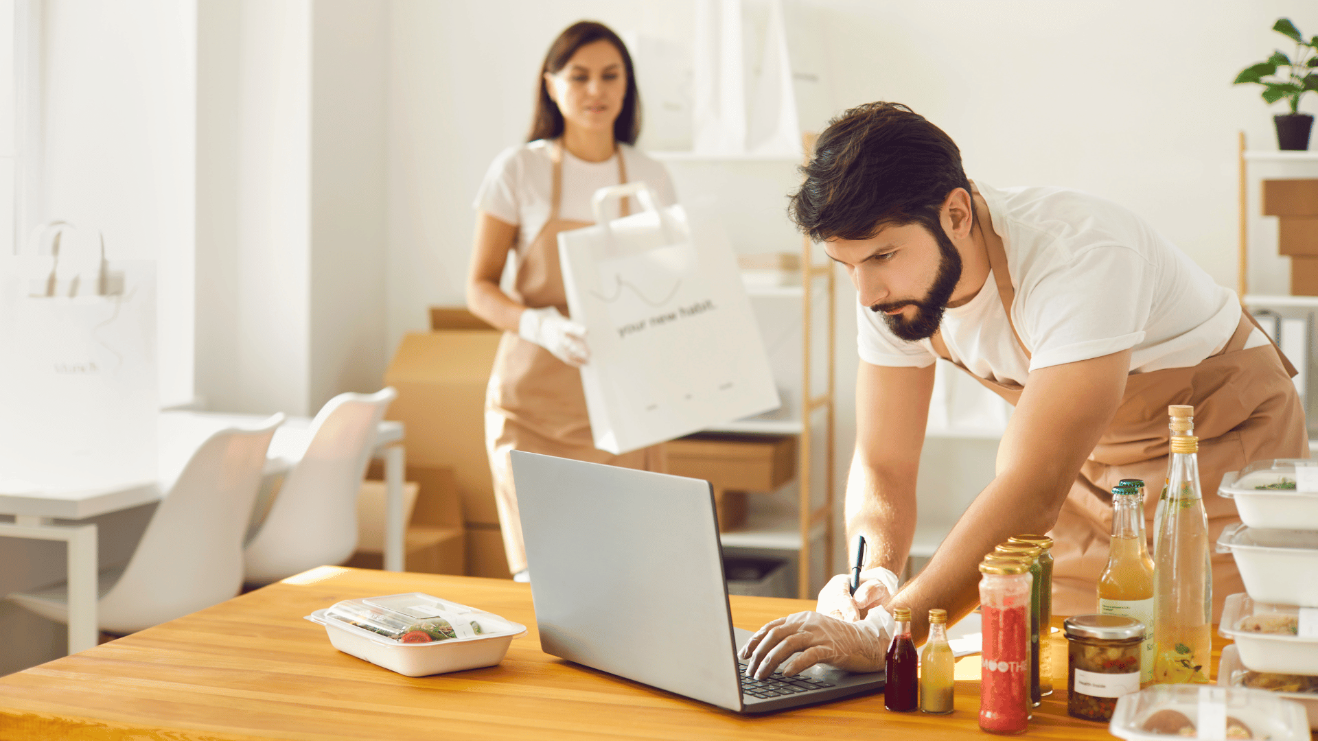 Man looking at computer with food around woman in background packing food into bag